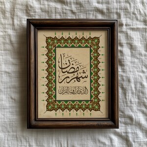 May include: A framed artwork featuring Arabic calligraphy in brown ink on a beige background. The frame is dark brown. The artwork is decorated with green and brown geometric patterns. The text in the artwork is in a stylized script.