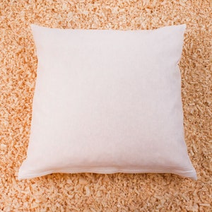May include: A white pillow with a smooth, plain surface. The pillow is lying on a bed of wood shavings.