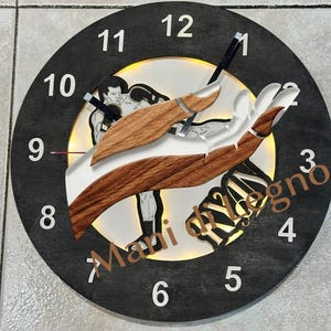 Può includere: Un orologio da parete circolare con una cornice grigio scuro testurizzata e numeri bianchi da 1 a 12. Il centro presenta un'illustrazione retroilluminata di un pugile in posizione di combattimento, sovrapposta a una scultura in legno e lancette.