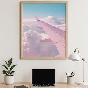 May include: A framed print of an airplane wing against a backdrop of clouds and sky. The wing is a soft pink, contrasting with the blue sky and fluffy white clouds. The print hangs above a desk with a laptop, plant, and desk lamp.
