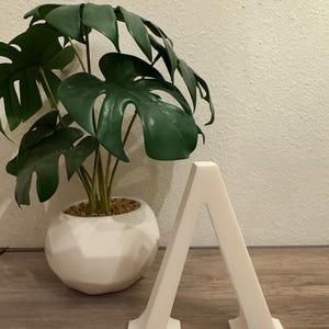 May include: A white, geometric letter "A" stands on a wooden surface. A potted plant with large, green leaves is in the background. The plant is in a white, spherical pot. The background is a light-colored wall.