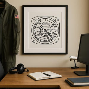 May include: Black and white framed print of an aircraft airspeed indicator. The gauge displays numbers and text, including "AIPHLI" and "A15SPEED". The print is displayed on a wooden desk with other items.