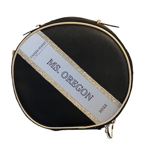 May include: Black round handbag with a gold zipper and trim. A white diagonal stripe is adorned with rhinestones and the text "MS. OREGON 2022" and "UNITED STATES of AMERICA".