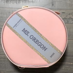 Op de afbeelding: Een ronde roze make-up tas met een zilveren en gouden lint waarop "UNITED STATES of AMERICA" en "MS. OREGON 2022" staat.