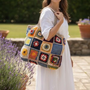 May include: A colorful, handmade crochet tote bag with a patchwork design. The bag features squares in various colors, including blue, yellow, red, and green. The bag has a neutral-colored strap and is held by a person wearing a white dress.
