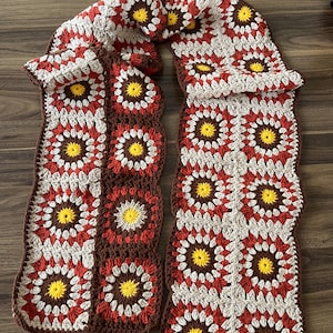 May include: A long scarf constructed from multiple crocheted granny squares. Each square features a bright yellow centre surrounded by concentric rings of dark brown, rust red, and cream-coloured yarn, creating a repeating floral-like pattern.