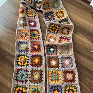 May include: A handmade crochet scarf with a patchwork design. The scarf features colourful squares in shades of orange, yellow, green, blue, and red, with a neutral border. The scarf is displayed on a wooden surface.