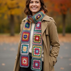 May include: A vibrant, hand-crocheted scarf with a granny square pattern. The scarf showcases squares in diverse colours, such as red, blue, yellow, and green, joined by a grey border. The scarf is worn over a white sweater and a tan trench coat.