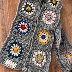 Puede incluir: Una bufanda de ganchillo gris con motivos florales coloridos. La bufanda está hecha de un hilo suave y texturizado y tiene cuadrados de flores de ganchillo en tonos de azul, rojo, amarillo y rosa. Se adjunta una pequeña etiqueta con texto.