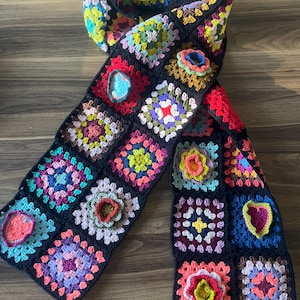 Könnte beinhalten: Ein langer, bunter Häkel-Schal aus Granny Squares in verschiedenen Farben, darunter Rot, Grün, Blau, Pink und Gelb. Der Schal ist aus schwarzem Garn mit bunten Quadraten.