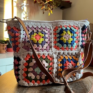 May include: A crocheted bag with a brown leather strap. The bag is made of granny squares in various colors, including pink, orange, green, blue, and yellow.