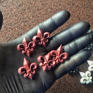 Könnte beinhalten: Eine Sammlung kleiner, dreidimensionaler Fleur-de-Lis-Embleme. Vier dieser Ornamente sind dunkelrot und liegen auf einer Handfläche, während mehrere andere in Weiß und Dunkelgrau in einer Reihe auf einer strukturierten Oberfläche liegen.