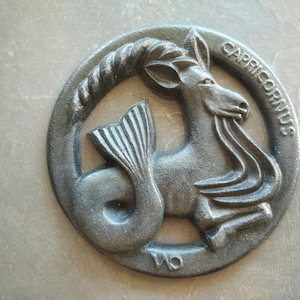 May include: Black cast iron zodiac sign of Capricorn. The sign is a circular shape with a detailed depiction of the sea-goat, with the words "Capricornus" inscribed around the edge.