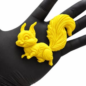 Pode incluir: Uma estatueta de esquilo amarelo brilhante com uma cauda felpuda detalhada e pelo texturizado. A pequena figura animal moldada é representada numa posição sentada com orelhas pontiagudas e olhos grandes, exibindo uma cor sólida e vibrante.
