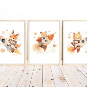 May include: Set of three framed watercolor art prints featuring foxes in space. Each print shows a fox astronaut riding a rocket ship, surrounded by stars and planets. The artwork has a soft, whimsical style with warm colors.