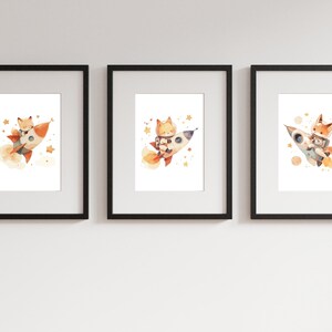 May include: Set of three framed art prints featuring watercolor illustrations of a fox in a rocket ship. The fox is depicted in various poses, surrounded by stars and planets. The prints are framed in black.