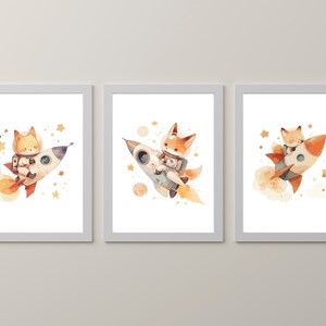 May include: Set of three framed watercolor prints featuring a fox astronaut in a rocket ship. The illustrations have a whimsical style with orange, white, and blue rockets, surrounded by stars and planets. The artwork is set against a white background.