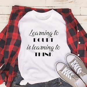 May include: White t-shirt with black text that reads "Learning to Doubt is learning to Think". The shirt is laid out on a wooden surface with a red and black plaid shirt and a pair of gray sneakers.