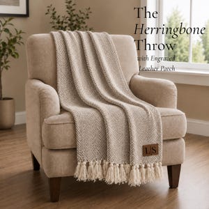 May include: A beige and white herringbone throw blanket with fringe tassels at the bottom edge. The blanket features a rectangular brown leather patch in the lower corner with the engraved letters LS. The text The Herringbone Throw with Engraved Leather Patch is visible.