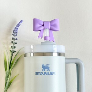 Pu&ograve; includere: Una tazza da viaggio Stanley bianca con un manico grigio e un logo blu chiaro che dice "STANLEY". La tazza ha un coperchio bianco con un fiocco viola sopra la cannuccia.