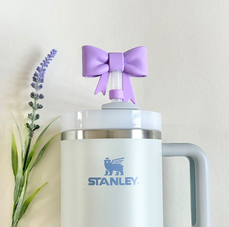 Pu&ograve; includere: Una tazza da viaggio Stanley bianca con un manico grigio e un logo blu chiaro che dice "STANLEY". La tazza ha un coperchio bianco con un fiocco viola sopra la cannuccia.
