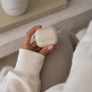 K&ouml;nnte beinhalten: Eine beige Silikon-Schutzh&uuml;lle f&uuml;r kabellose Ohrh&ouml;rer mit dem eingepr&auml;gten Namen Sophia in Schreibschrift auf der Vorderseite. Die H&uuml;lle hat eine glatte, matte Oberfl&auml;che und eine kleine seitliche Schlaufe, passend f&uuml;r g&auml;ngige Ladeetuis von Ohrh&ouml;rern.