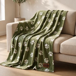 May include: An olive green fleece blanket featuring a repeating pattern of white paw prints. The blanket is draped over a sofa and includes a small, rectangular brown leather-like patch in the bottom corner with the name Bella embossed on it.