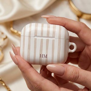 May include: A white and beige striped protective case for wireless earbuds featuring a small circular loop on the right side. The front of the case is personalized with the dark brown capital letters HM centered near the bottom edge.