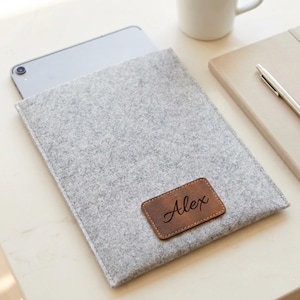 Op de afbeelding: Een grijze vilten tablethoes met een bruin leren label met de tekst "Alex" in sierlijke letters. De hoes bevat een zilveren tablet en ligt op een lichtgekleurd oppervlak.