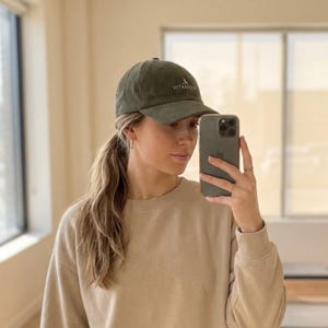 May include: Olive green baseball cap with the word "VITAMIN" embroidered on the front. The cap has a curved brim and a classic six-panel design. The hat is made of a soft, textured fabric.
