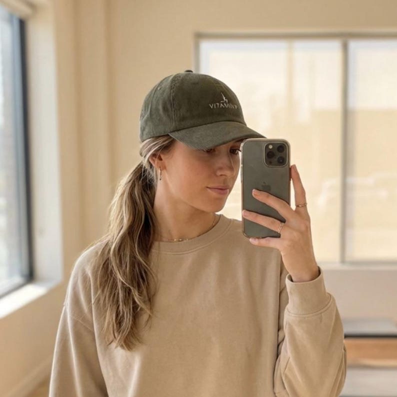 May include: Olive green baseball cap with the word "VITAMIN" embroidered on the front. The cap has a curved brim and a classic six-panel design. The hat is made of a soft, textured fabric.