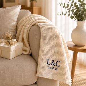 Peut inclure: Un plaid en tricot gaufré de couleur crème avec une broderie bleu foncé indiquant L&C et la date 26.07.26. Le plaid est drapé sur un canapé à côté d'une boîte cadeau blanche nouée avec un ruban et un petit bouquet de fleurs séchées.