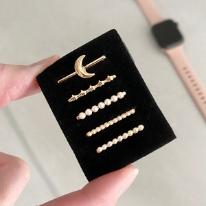 May include: A set of five gold-toned jewelry pieces on a black velvet background. The pieces include a crescent moon charm, a row of stars, and three rows of pearls.
