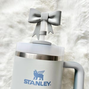 Pu&ograve; includere: Una tazza da viaggio Stanley bianca con un coperchio a forma di fiocco grigio e un logo di orso blu con la scritta "STANLEY" sotto.