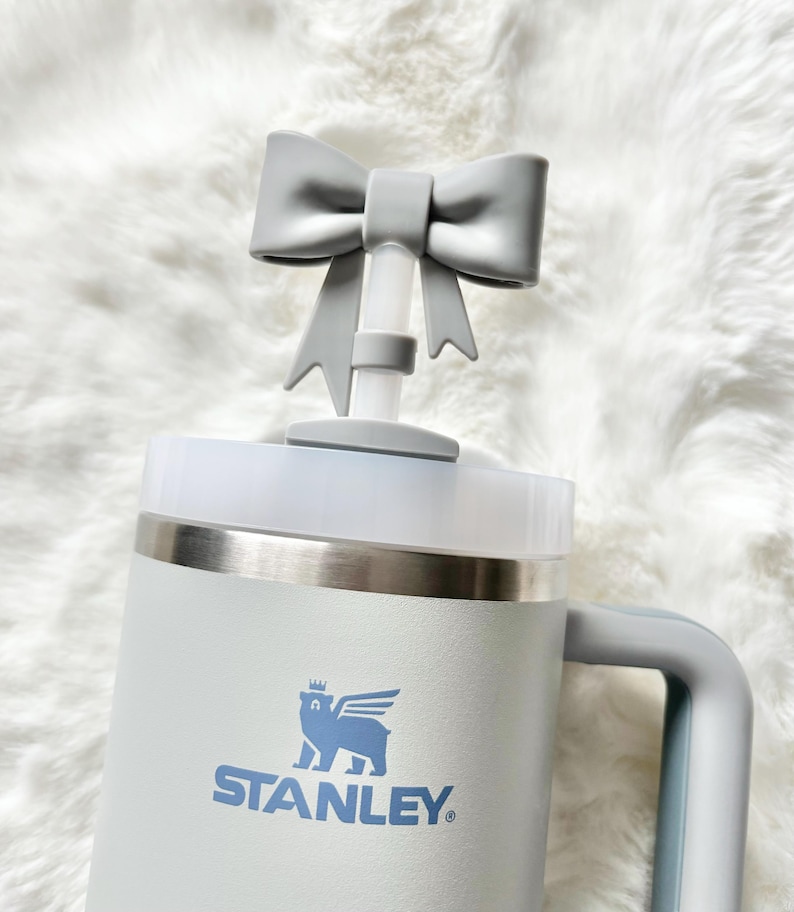 Pu&ograve; includere: Una tazza da viaggio Stanley bianca con un coperchio a forma di fiocco grigio e un logo di orso blu con la scritta "STANLEY" sotto.