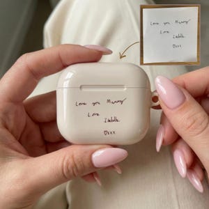 Könnte beinhalten: Ein glattes, cremefarbenes Ladecase für kabellose Ohrhörer mit einer individuellen schwarzen Handschrift, die lautet: Love you Mummy, Love, Isabelle, inklusive Herzsymbol und xxx. Ein kleiner Einschub zeigt die originale handschriftliche Notiz auf weißem Papier.