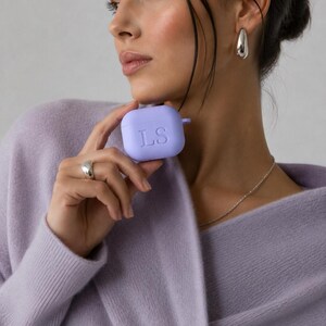 K&ouml;nnte beinhalten: Eine hellviolette Schutzh&uuml;lle aus Silikon f&uuml;r kabellose Ohrh&ouml;rer mit den eingepr&auml;gten Buchstaben LS auf der Vorderseite. Die H&uuml;lle hat eine glatte, matte Oberfl&auml;che und eine kleine seitliche Schlaufe in einem einfarbigen Lavendelton.