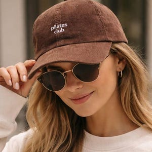 May include: A brown corduroy baseball cap with the text "pilates club" embroidered in white. The cap has a curved brim and is worn by a person wearing sunglasses.