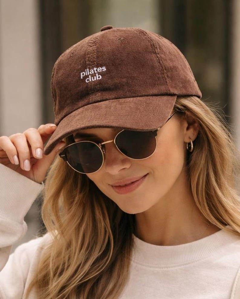 May include: A brown corduroy baseball cap with the text "pilates club" embroidered in white. The cap has a curved brim and is worn by a person wearing sunglasses.