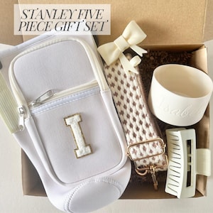 Może przedstawiać: Biały zestaw upominkowy w kartonowym pudełku zawiera torebkę crossbody ze złotą literą "I", pasek we wzory, spinkę do włosów, kokardę i mały kubek z wygrawerowanym imieniem "Isabel". U góry znajduje się napis "STANLEY FIVE PIECE GIFT SET".