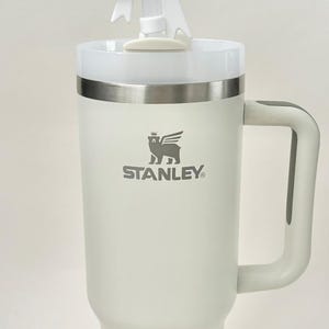Pu&ograve; includere: Una tazza da viaggio Stanley bianca con un coperchio in acciaio inossidabile e un fondo in gomma bianca. La tazza ha un logo grigio con la scritta "STANLEY" e un leone che indossa una corona sopra il testo. Un fiocco bianco &egrave; posto sulla parte superiore della tazza.