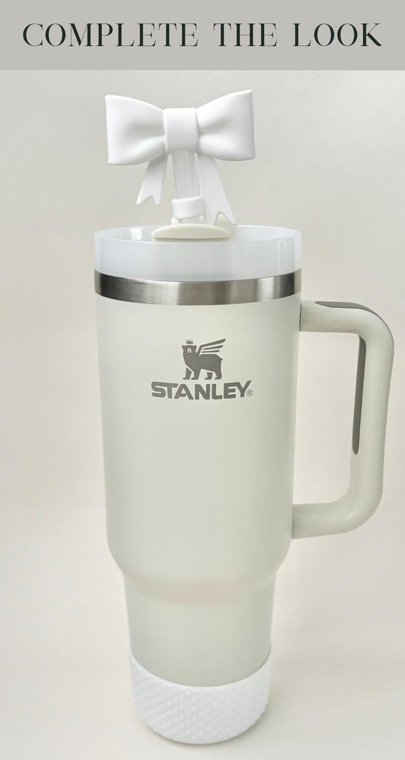 Pu&ograve; includere: Una tazza da viaggio Stanley bianca con un coperchio in acciaio inossidabile e un fondo in gomma bianca. La tazza ha un logo grigio con la scritta "STANLEY" e un leone che indossa una corona sopra il testo. Un fiocco bianco &egrave; posto sulla parte superiore della tazza.