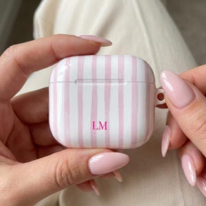 Könnte beinhalten: Weißes AirPod-Etui mit rosa vertikalen Streifen und den Initialen "LM" in Rosa. Das Etui wird von einer Person mit langen, rosa Nägeln gehalten. Das Etui hat eine abgerundete quadratische Form.
