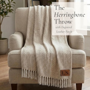 May include: A beige herringbone throw blanket with fringe, draped over a beige armchair. The throw has a leather patch with the initials "LS". The text on the image reads "The Herringbone Throw with Engraved Leather Patch".