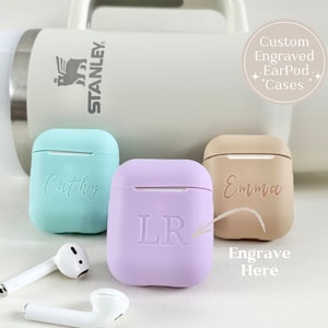 Pode incluir: Três capas para AirPod personalizadas, uma azul claro com o nome "Cathy" gravado, uma roxo claro com as iniciais "LR" gravadas e uma marrom claro com o nome "Emma" gravado. As capas estão sobre uma superfície branca com dois fones de ouvido AirPod brancos.