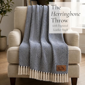 May include: A navy blue and white herringbone patterned throw blanket with cream-coloured tassels along the bottom edge. A rectangular brown leather patch with the engraved letters LS is attached to the lower right corner. Text reads The Herringbone Throw with Engraved Leather Patch.