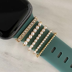 May include: A teal green Apple Watch band with four gold and pearl embellishments. The embellishments are a row of small pearls, a row of larger pearls, a row of smaller pearls, and a row of rhinestones.