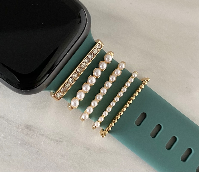 May include: A teal green Apple Watch band with four gold and pearl embellishments. The embellishments are a row of small pearls, a row of larger pearls, a row of smaller pearls, and a row of rhinestones.