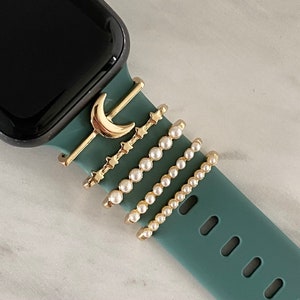May include: A teal green Apple Watch band with five gold-toned charms attached. The charms include a crescent moon, a row of stars, and three rows of pearls.