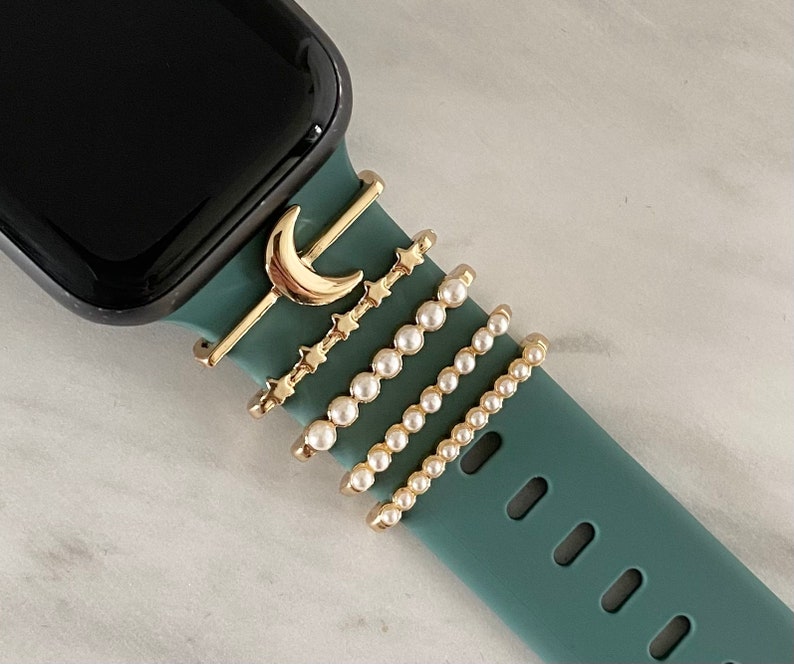 May include: A teal green Apple Watch band with five gold-toned charms attached. The charms include a crescent moon, a row of stars, and three rows of pearls.